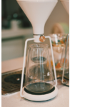 A white funnel narrowing into a glass vessel, symbolizing abundant information being filtered into scarce human attention in the AI economy.