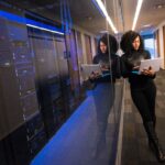 Data center professional walking beside server racks with a tablet, representing AI infrastructure and the changing workforce.