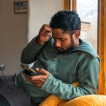 A man sitting by a window looks confused and stressed while scrolling through his smartphone, illustrating subscription fatigue and budget management.