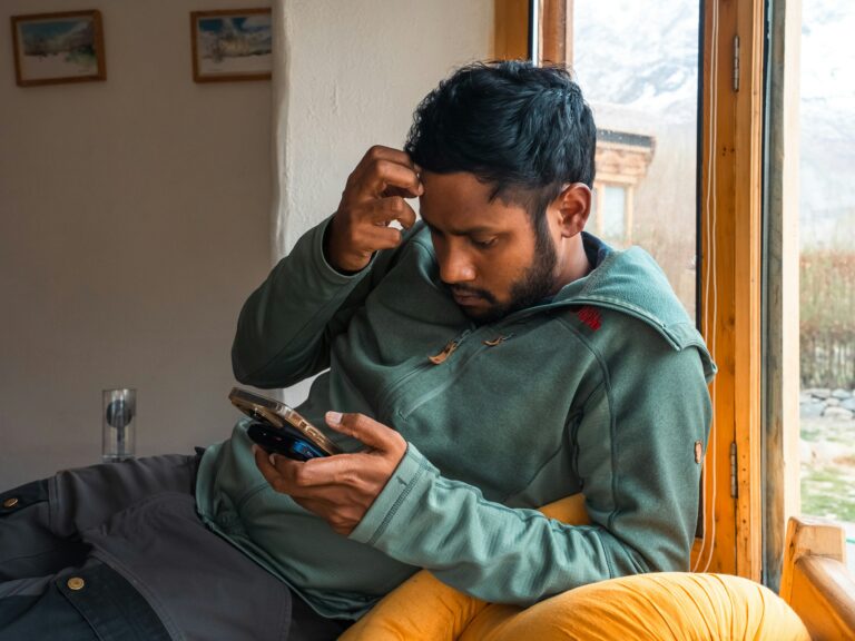 A man sitting by a window looks confused and stressed while scrolling through his smartphone, illustrating subscription fatigue and budget management.