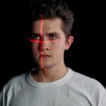 A young man with a red laser crosshair projected onto his face against a black background, illustrating facial recognition, identity verification, and biometric surveillance technology.