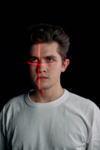 A young man with a red laser crosshair projected onto his face against a black background, illustrating facial recognition, identity verification, and biometric surveillance technology.