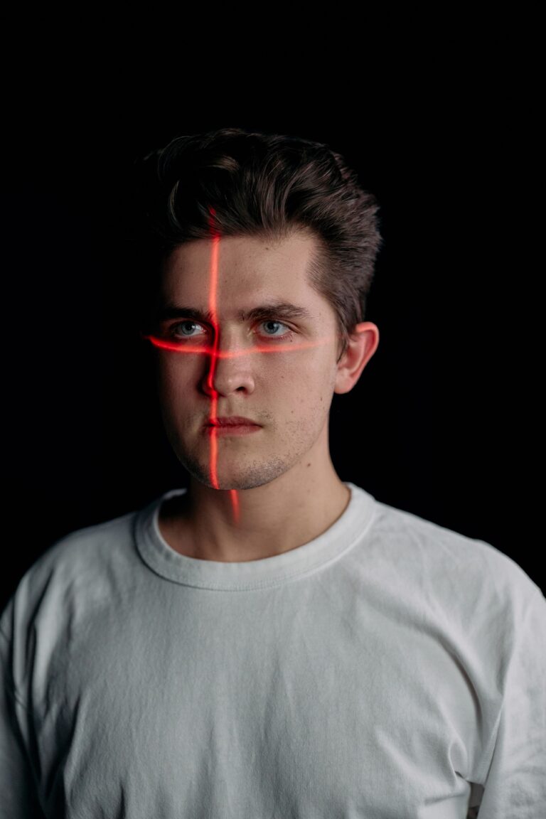A young man with a red laser crosshair projected onto his face against a black background, illustrating facial recognition, identity verification, and biometric surveillance technology.