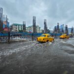 Yellow taxis driving through deeply flooded city streets next to concrete metro construction pillars during a severe monsoon downpour in India.