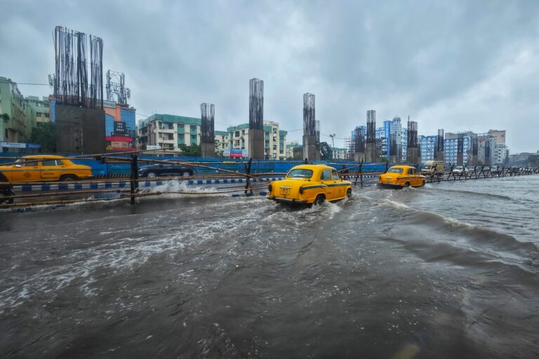 Yellow taxis driving through deeply flooded city streets next to concrete metro construction pillars during a severe monsoon downpour in India.