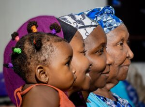 Four generations of a family shown in profile, from a young child to a great-grandmother, wearing colorful traditional headwraps