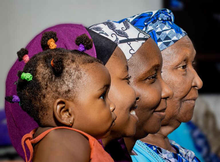 Four generations of a family shown in profile, from a young child to a great-grandmother, wearing colorful traditional headwraps