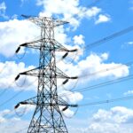 A high-voltage electricity transmission tower carrying power lines against a bright blue sky with white clouds, representing modern power grid infrastructure.
