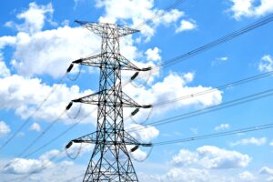 A high-voltage electricity transmission tower carrying power lines against a bright blue sky with white clouds, representing modern power grid infrastructure.
