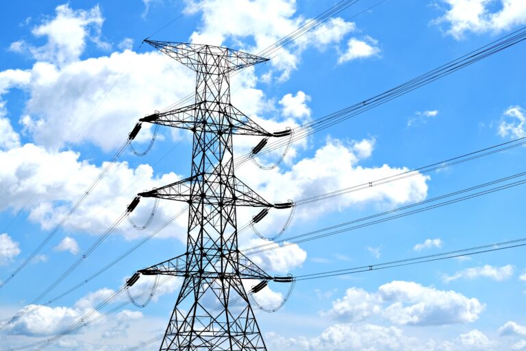 A high-voltage electricity transmission tower carrying power lines against a bright blue sky with white clouds, representing modern power grid infrastructure.