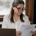 Older female professional with glasses reviewing financial documents at a wooden desk with a laptop