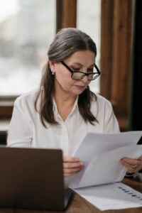 Older female professional with glasses reviewing financial documents at a wooden desk with a laptop