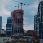An unfinished high-rise building lit orange by construction lighting at dusk, wrapped in cranes and boxed in by completed glass towers on a city skyline