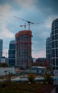 An unfinished high-rise building lit orange by construction lighting at dusk, wrapped in cranes and boxed in by completed glass towers on a city skyline