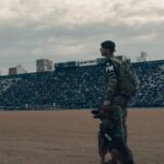 Military police officer with a security dog standing inside a stadium as a large crowd watches from the stands.
