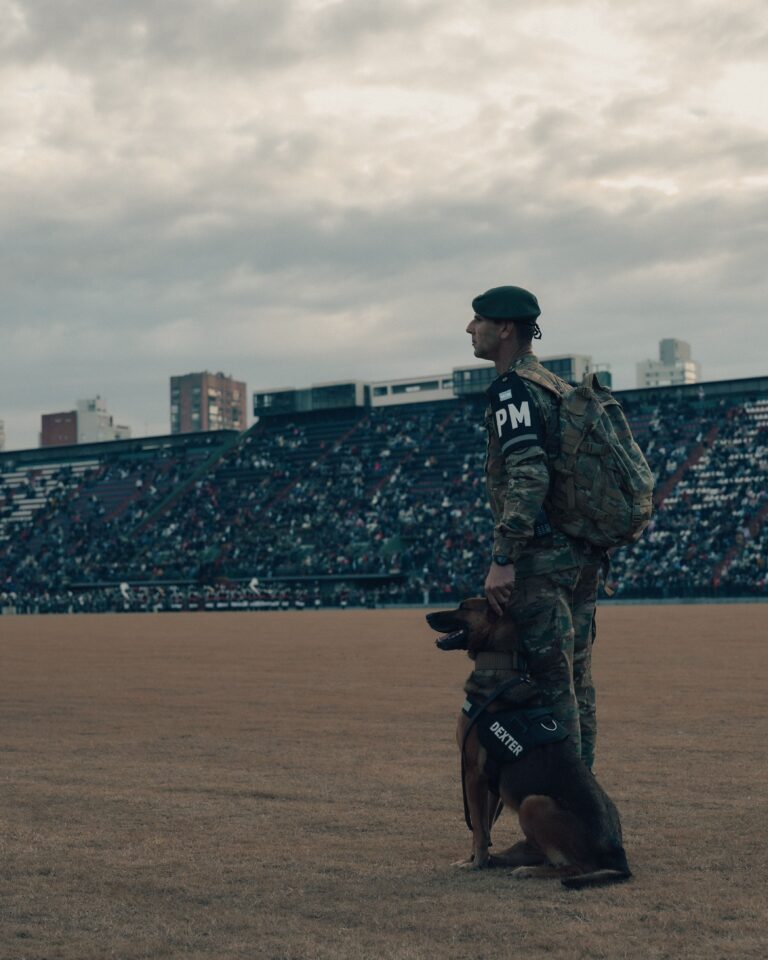 Military police officer with a security dog standing inside a stadium as a large crowd watches from the stands.