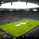 A packed football stadium before kickoff, with tens of thousands of supporters gathered around the pitch during a major international match.