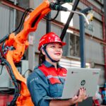 Industrial worker using a laptop beside a robotic arm in a smart factory, illustrating how AI-powered automation is transforming modern work and business operations.