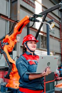 Industrial worker using a laptop beside a robotic arm in a smart factory, illustrating how AI-powered automation is transforming modern work and business operations.