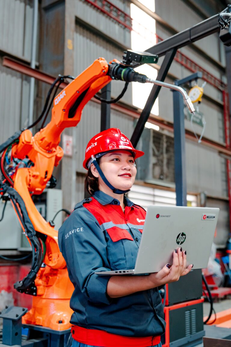 Industrial worker using a laptop beside a robotic arm in a smart factory, illustrating how AI-powered automation is transforming modern work and business operations.