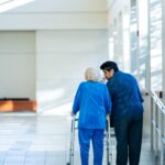A caregiver walking alongside an elderly person using a walker in a long term care facility, illustrating the growing demand for elder care.