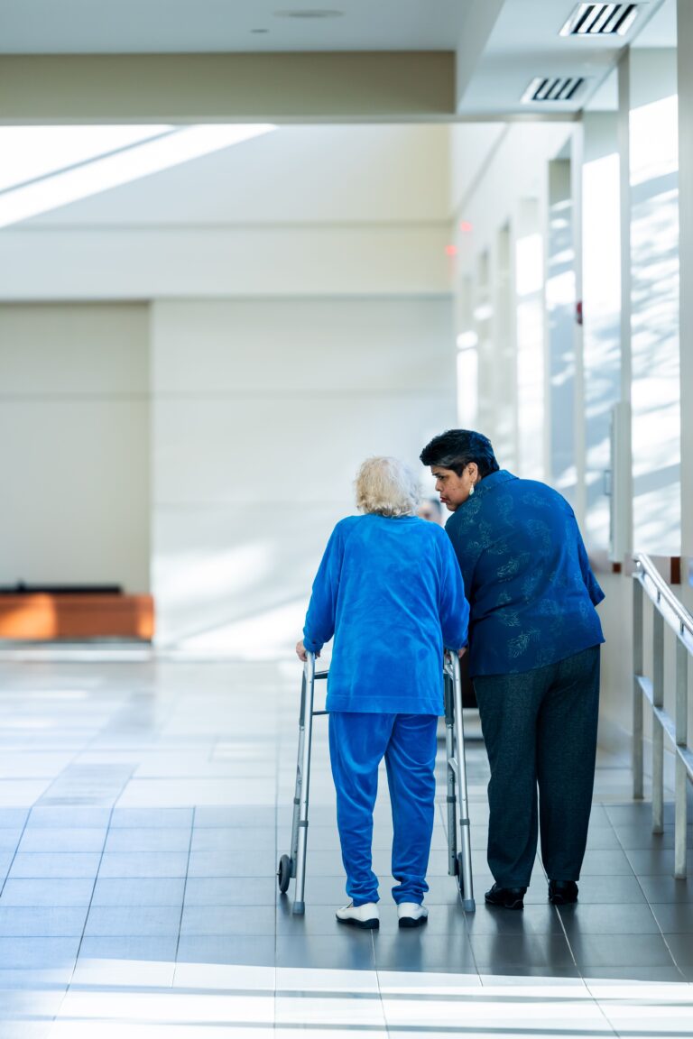A caregiver walking alongside an elderly person using a walker in a long term care facility, illustrating the growing demand for elder care.