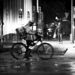 Gig economy delivery worker waiting with a bicycle on a wet city street at night