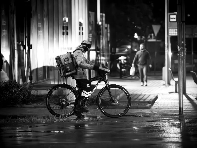 Gig economy delivery worker waiting with a bicycle on a wet city street at night