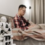 Small humanoid robot sitting beside a person in a bedroom, illustrating robots entering everyday home life