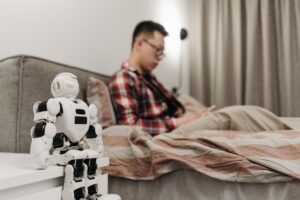 Small humanoid robot sitting beside a person in a bedroom, illustrating robots entering everyday home life
