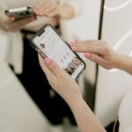 Person browsing a social media app on a smartphone while standing in front of a mirror, illustrating how algorithms influence fashion and personal identity.