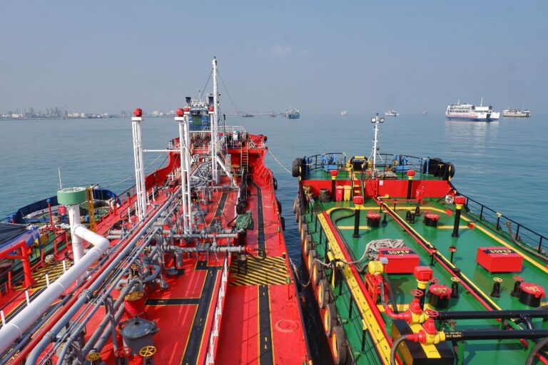 Two oil tankers positioned side by side at sea, with deck pipelines visible during a ship-to-ship transfer operation.