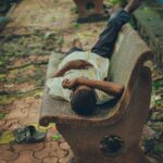 A man lying across a concrete bench in a public park, illustrating how people use urban spaces for rest.