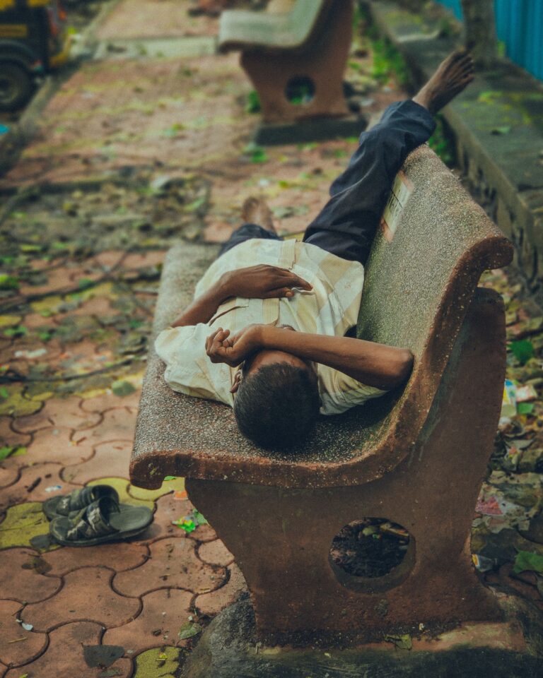 A man lying across a concrete bench in a public park, illustrating how people use urban spaces for rest.