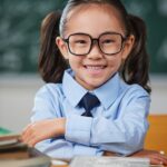 Schoolgirl wearing glasses seated at a desk with books in front of a classroom chalkboard.