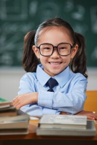 Schoolgirl wearing glasses seated at a desk with books in front of a classroom chalkboard.